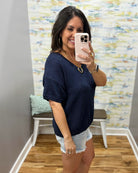 Woman taking a mirror selfie wearing a navy blue shirt and denim shorts in a room with patterned wallpaper.
