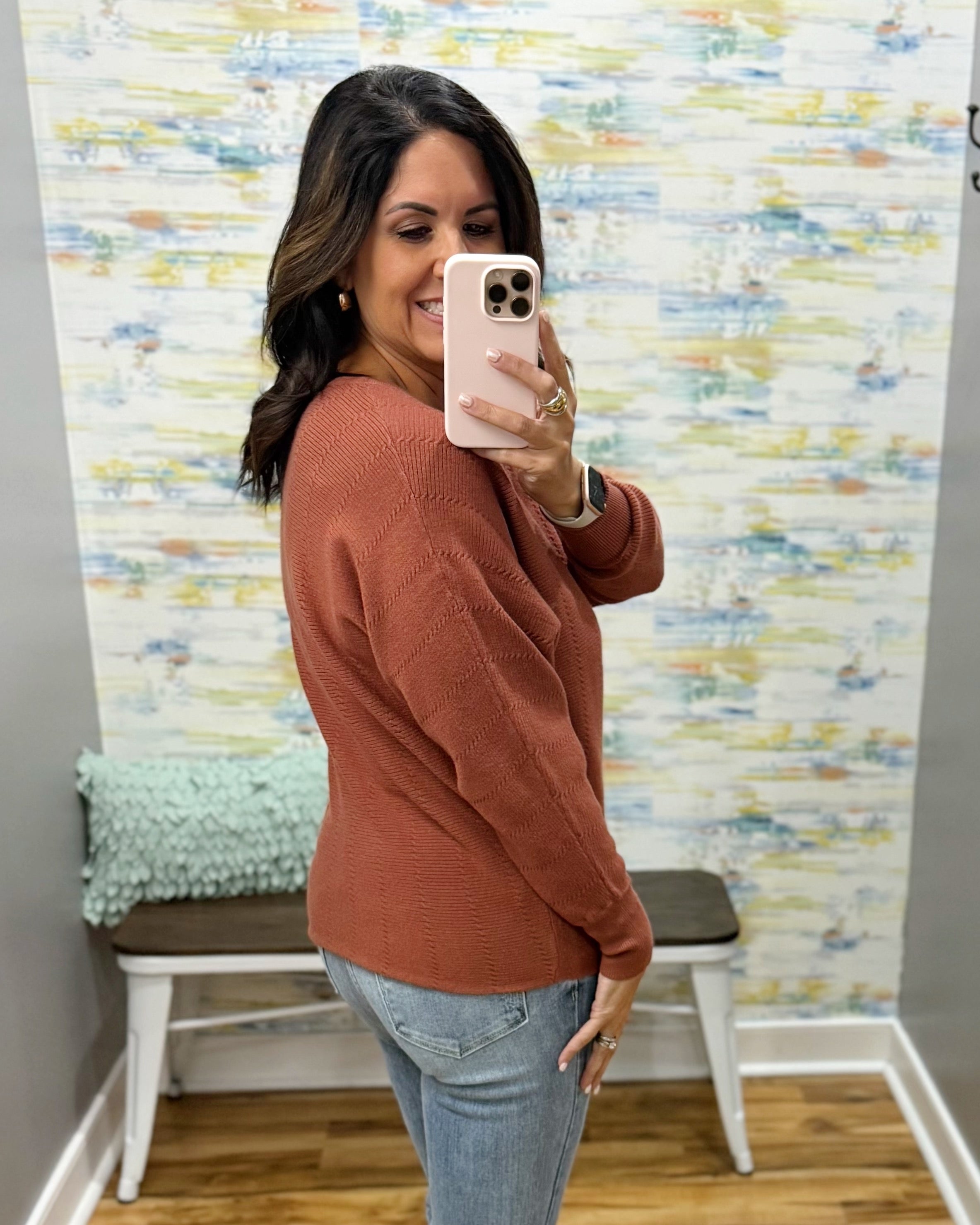 A woman takes a mirror selfie wearing a textured rust-orange long-sleeve knit sweater and light-wash blue jeans in a home setting with patterned wallpaper and a bench in the background.