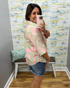 Woman taking a mirror selfie wearing a pink patterned shirt and denim shorts in a room with a colorful wall.