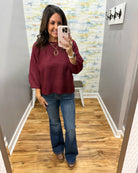 A woman takes a mirror selfie in a fitting room wearing a heathered burgundy long-sleeve top and blue flare jeans.