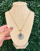 A gold chain necklace with a round gray stone pendant and a toggle clasp, displayed on a beige fabric jewelry bust. A hand with purple nail polish holds the left side of the bust, and a green leafy hedge serves as the background.