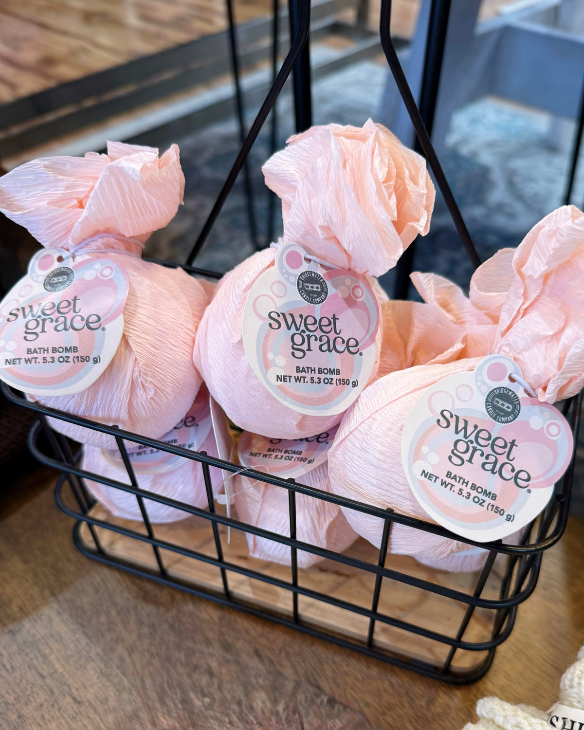 Sweet Grace pink bath bombs wrapped in tissue paper and attached with tags, displayed in a black wire basket on a wooden surface.