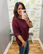 A woman taking a mirror selfie wearing a loose-fitting, short-sleeve knit sweater in deep burgundy and blue jeans, holding a light pink smartphone.