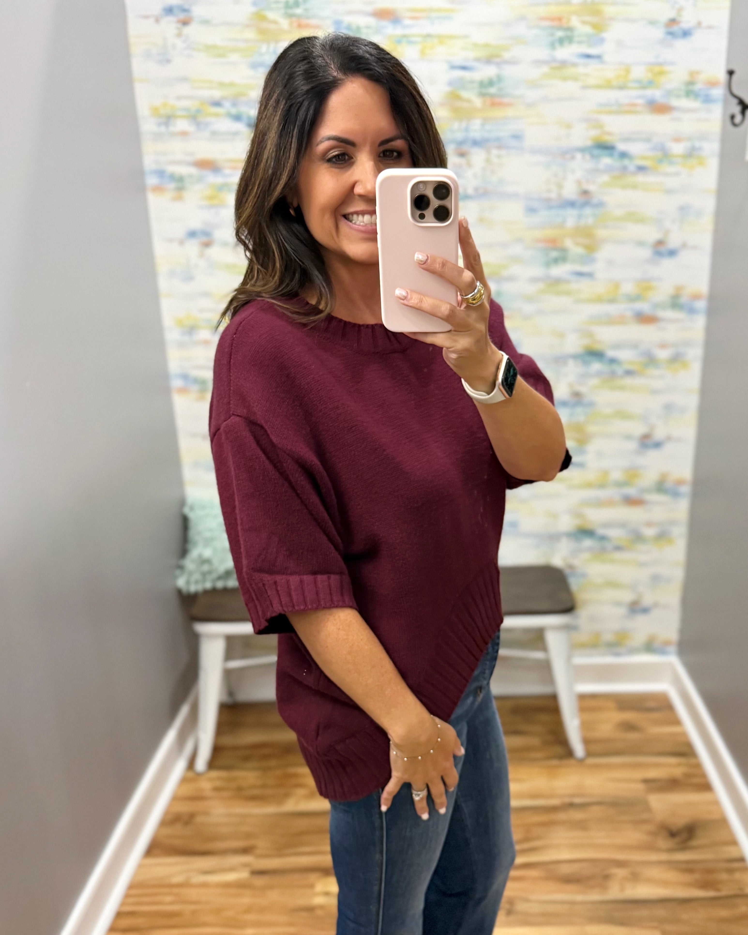 A woman taking a mirror selfie wearing a loose-fitting, short-sleeve knit sweater in deep burgundy and blue jeans, holding a light pink smartphone.
