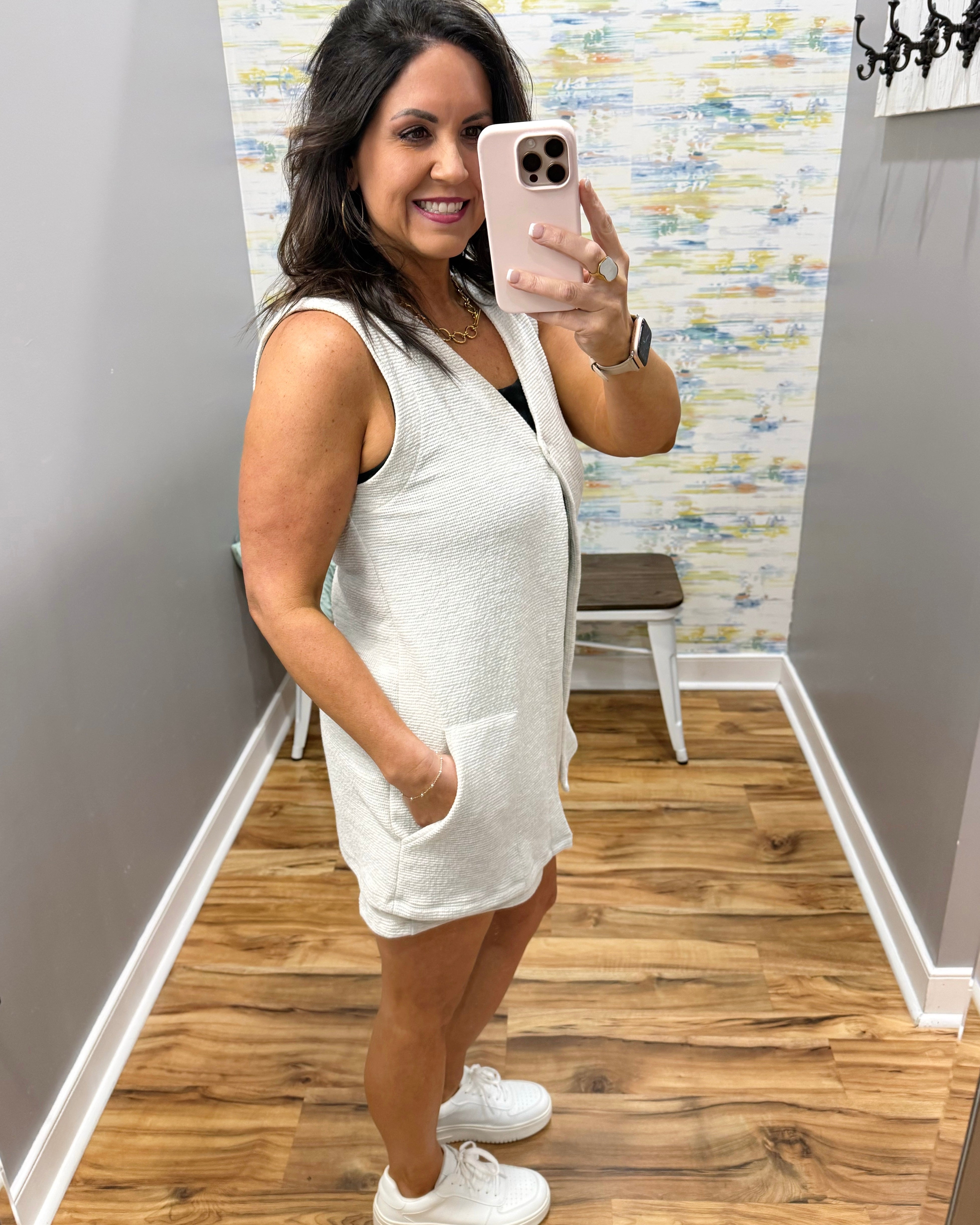 Woman taking a mirror selfie wearing a white sleeveless dress in a room with wooden flooring and patterned wallpaper.