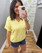 Woman taking a mirror selfie wearing a yellow shirt and denim shorts.