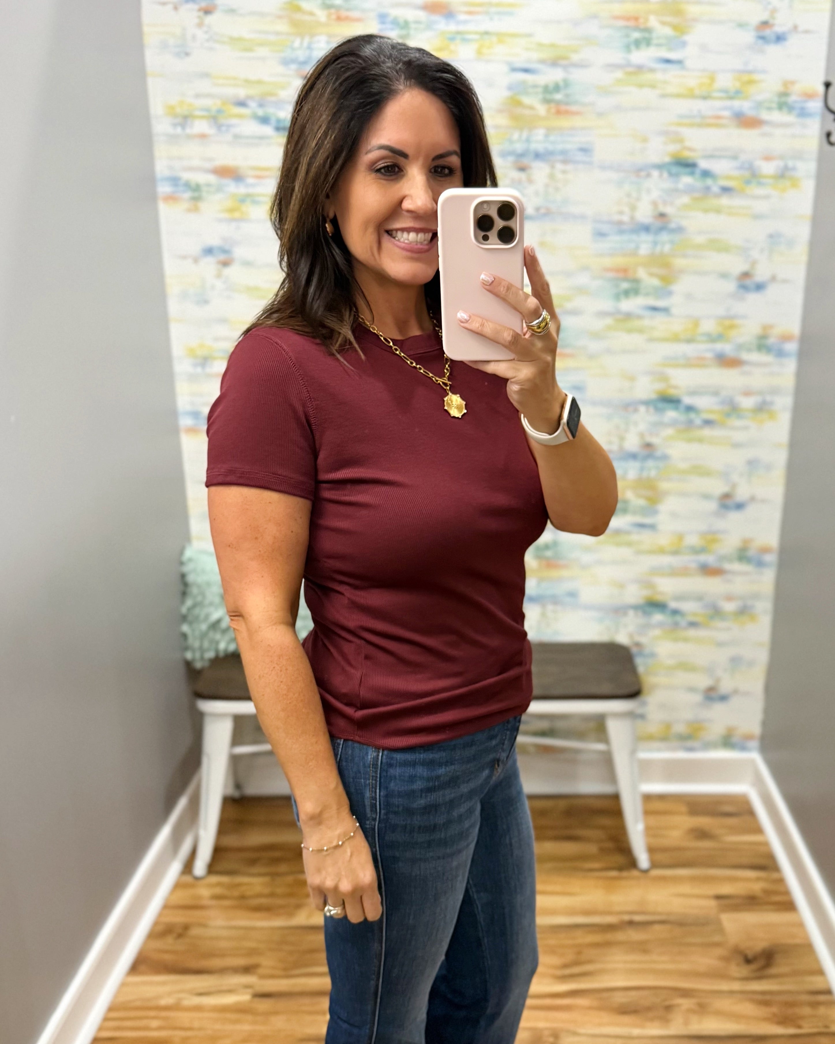 A woman takes a mirror selfie in a fitting room wearing a short-sleeve crewneck t-shirt in burgundy and blue jeans.