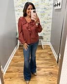 A woman takes a mirror selfie wearing a rust-colored ruffled blouse and dark blue flare jeans in a fitting room with wood floors and gray walls.