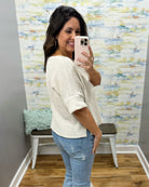 Woman taking a mirror selfie wearing a natural sweater and jeans in a room with a textured wall.