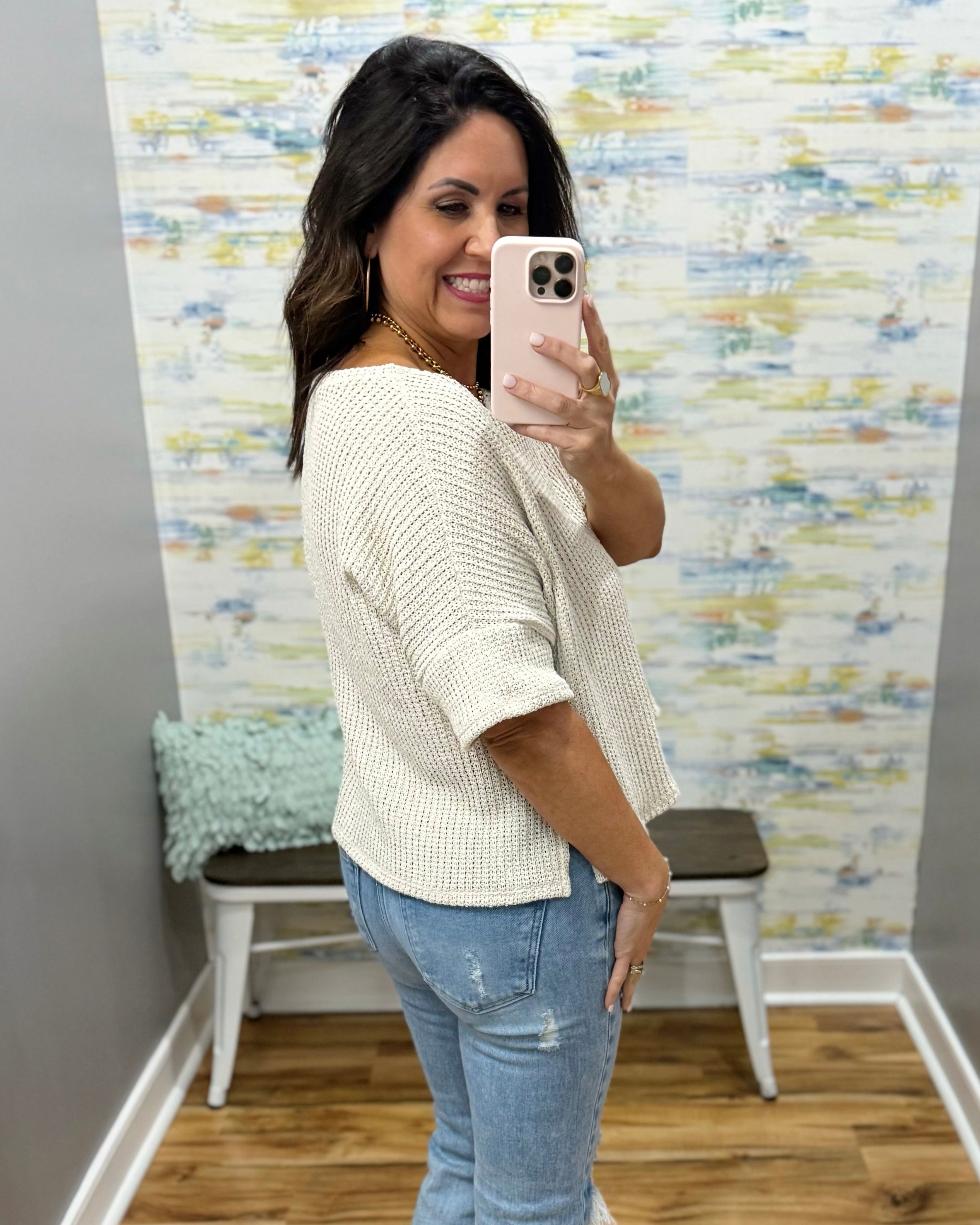 Woman taking a mirror selfie wearing a natural sweater and jeans in a room with a textured wall.