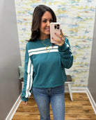A woman takes a mirror selfie wearing a teal long-sleeve sweatshirt with white stripes down the sleeves and across the chest, paired with blue jeans.