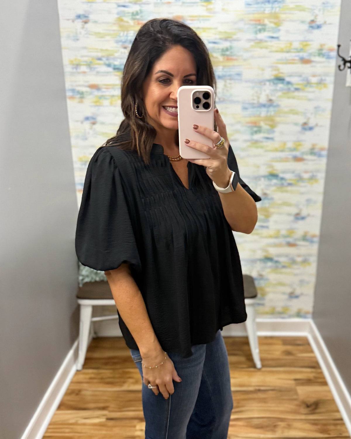 A woman takes a mirror selfie wearing a black short-sleeve blouse with pleated detailing and blue jeans in a boutique setting.