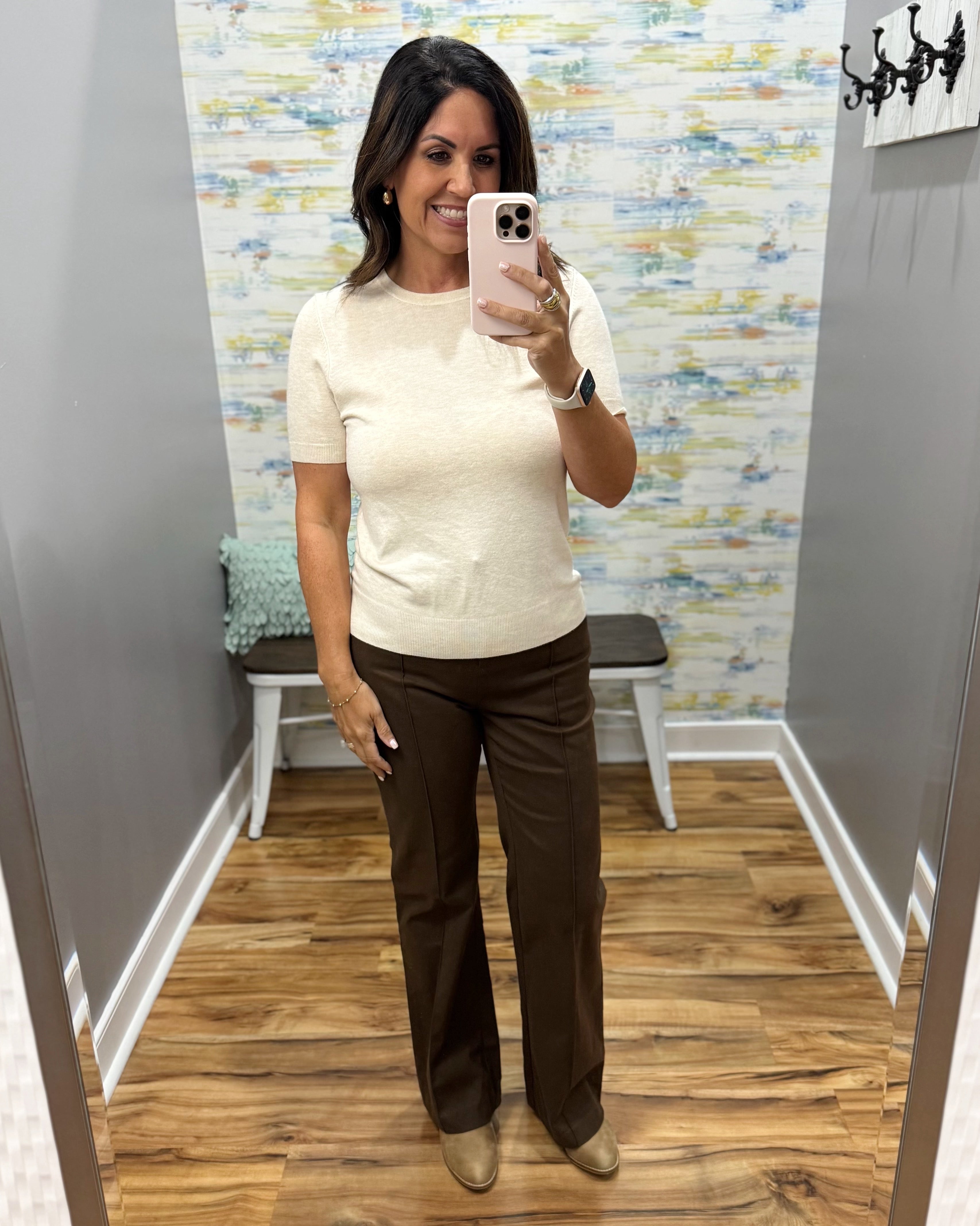 A woman takes a mirror selfie in a fitting room wearing a cream short-sleeve knit top and dark brown straight-leg pants.