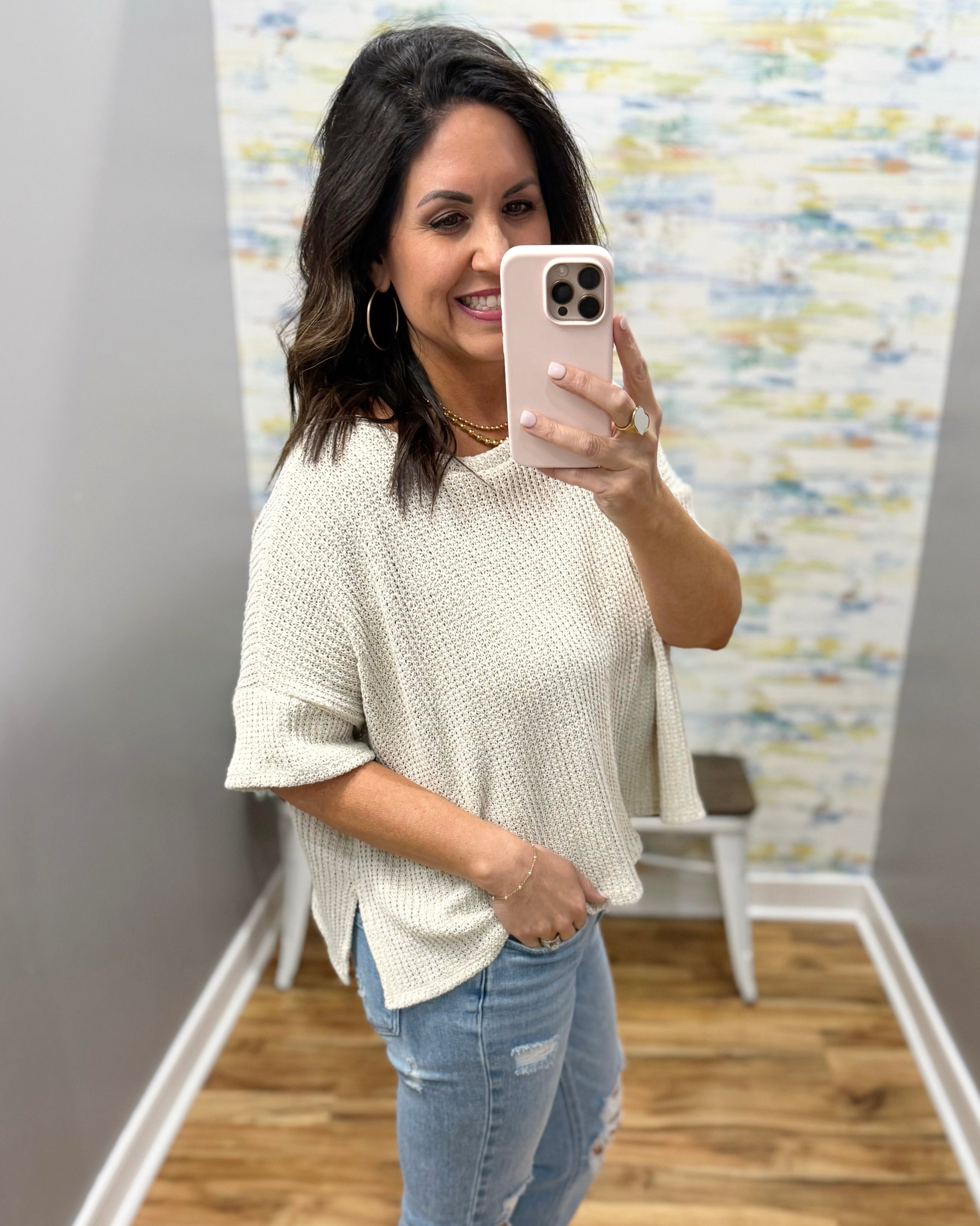 Woman taking a mirror selfie wearing a natural sweater and jeans in a room with a textured wall.
