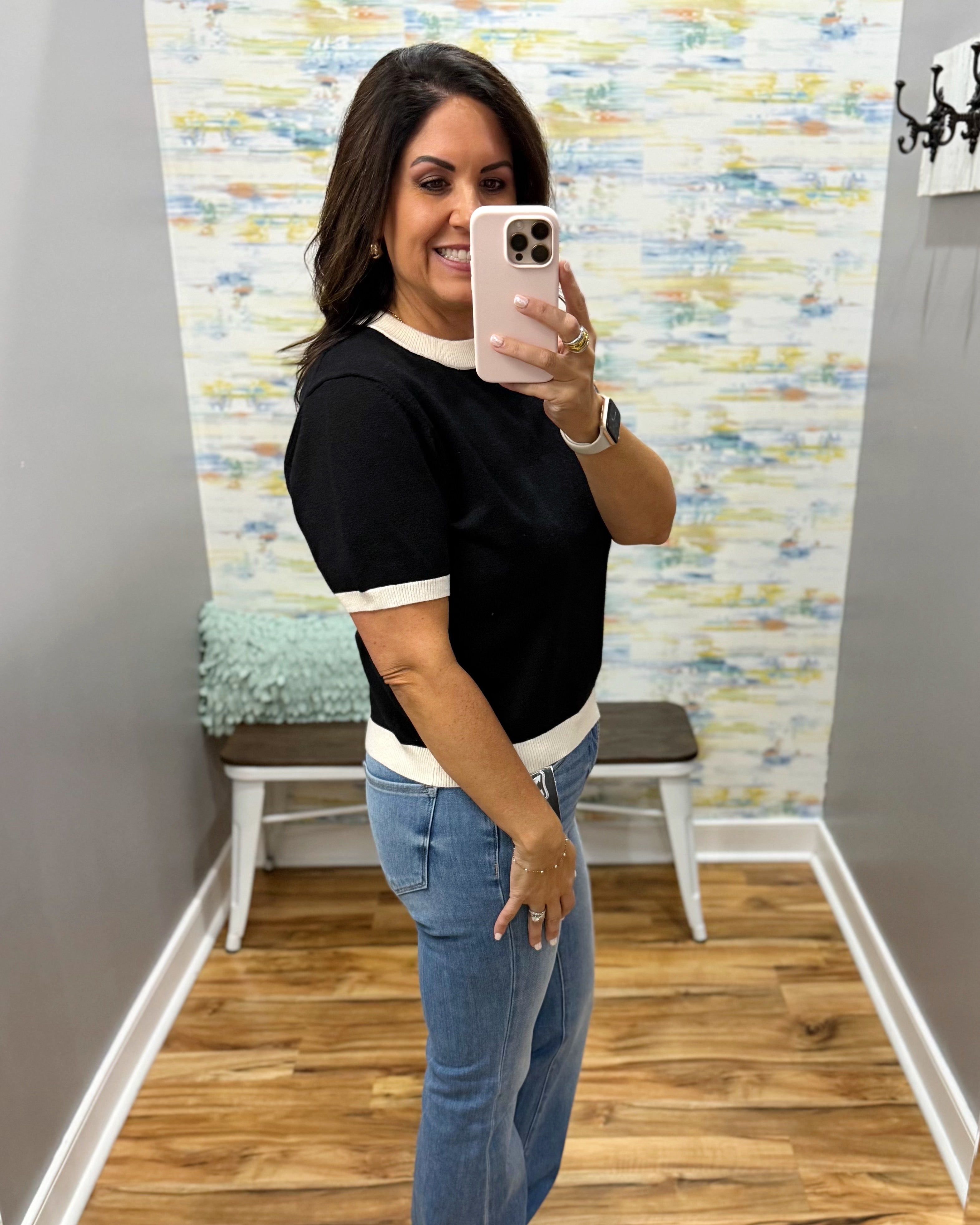 A woman takes a mirror selfie wearing a black short-sleeve t-shirt with white trim and blue jeans in a fitting room with a wood floor and abstract wallpaper.
