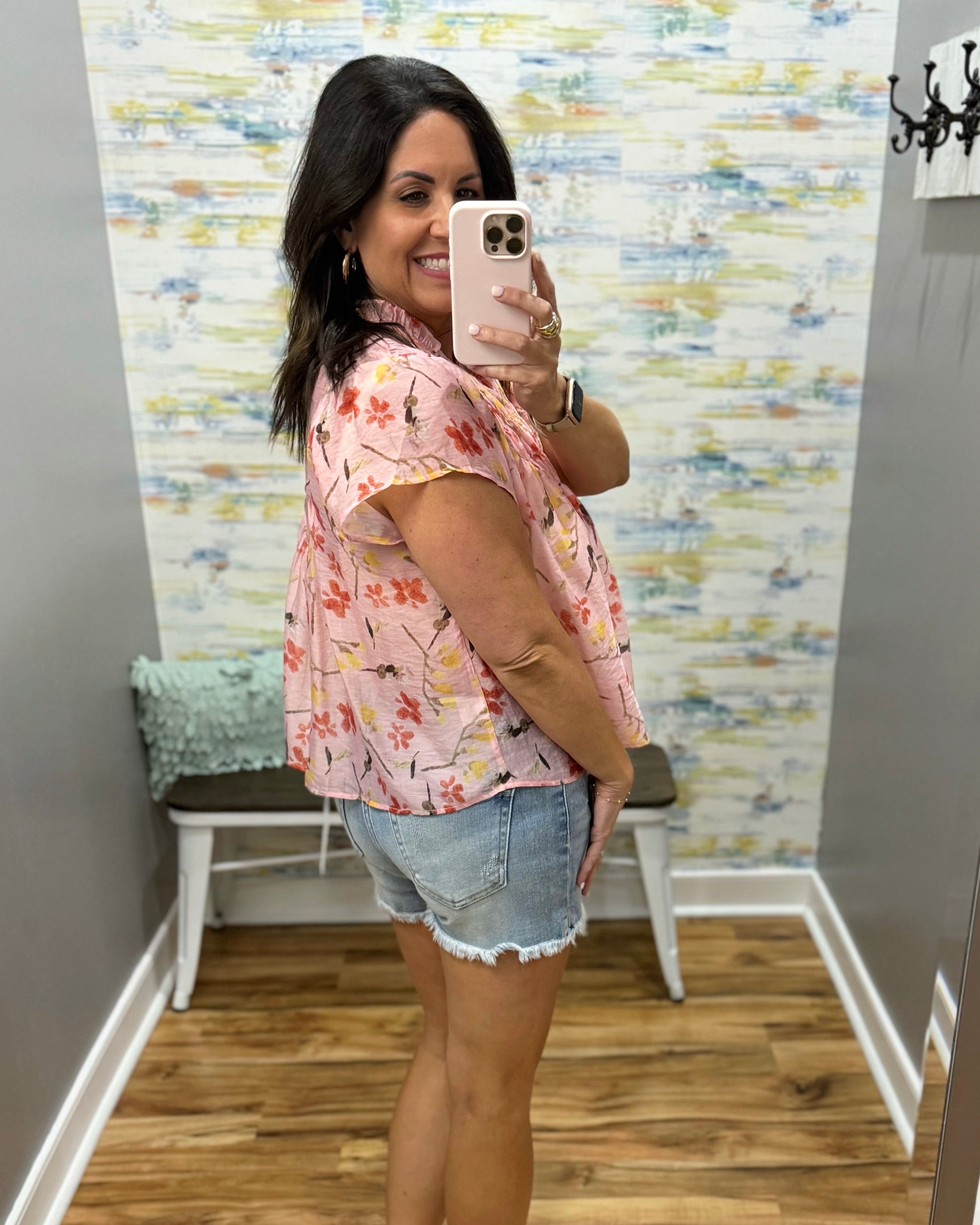 Woman taking a mirror selfie wearing a floral shirt and denim shorts in a room with colorful wall paper.