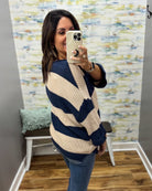 A woman takes a mirror selfie wearing a navy blue and cream striped knit sweater, blue jeans, and a gold ring, standing in a room with patterned wallpaper and a wooden floor.