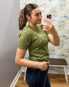 A woman takes a mirror selfie wearing a short-sleeve olive green knit sweater with small white embroidered dots, paired with dark blue jeans.