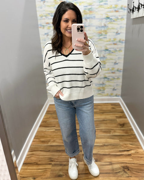 Woman taking a mirror selfie wearing a striped sweater and jeans in a room with patterned wallpaper.