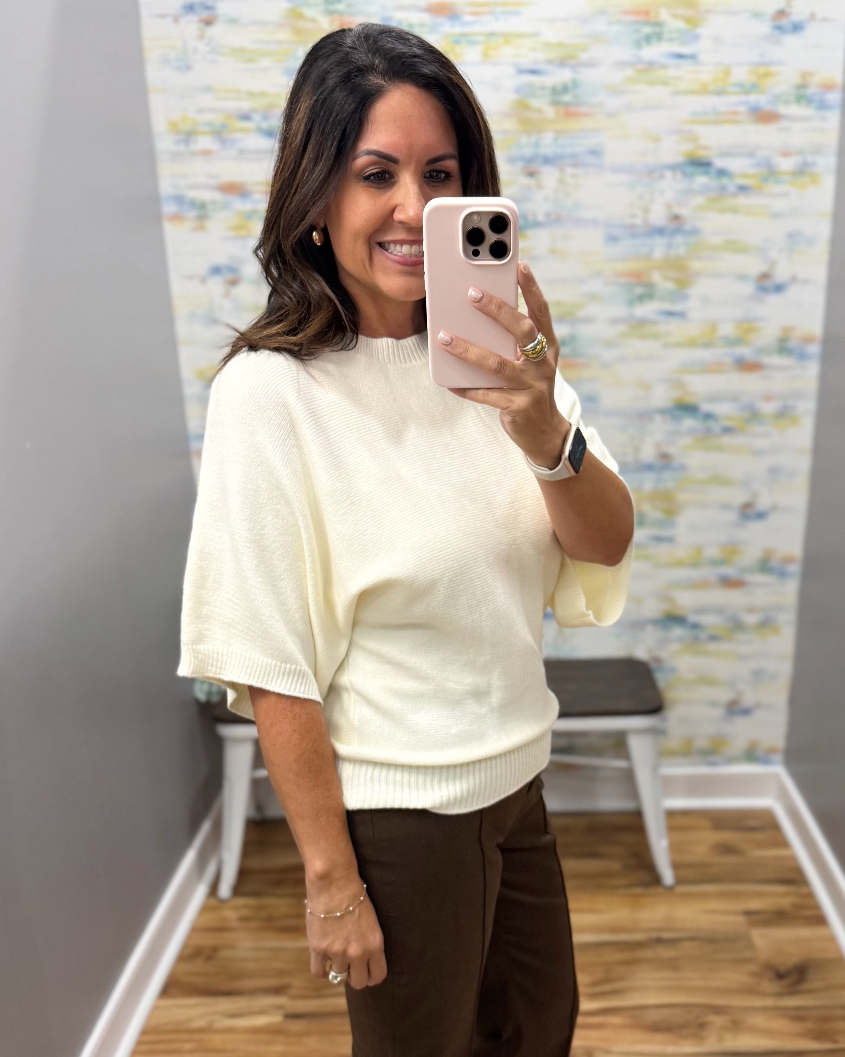 A woman taking a mirror selfie in a fitting room wearing a cream-colored short-sleeve knit sweater and dark brown pants.