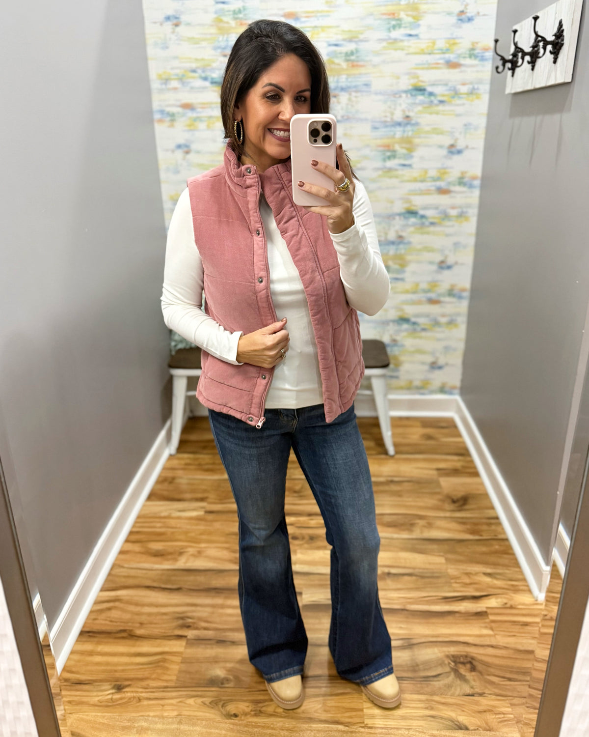 A woman takes a mirror selfie in a fitting room wearing a dusty pink sleeveless quilted vest over a white long-sleeve top, blue flare jeans, and tan boots.