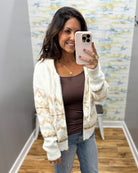A woman takes a mirror selfie wearing a cream and tan knit cardigan over a brown top and blue jeans, holding a light pink smartphone.