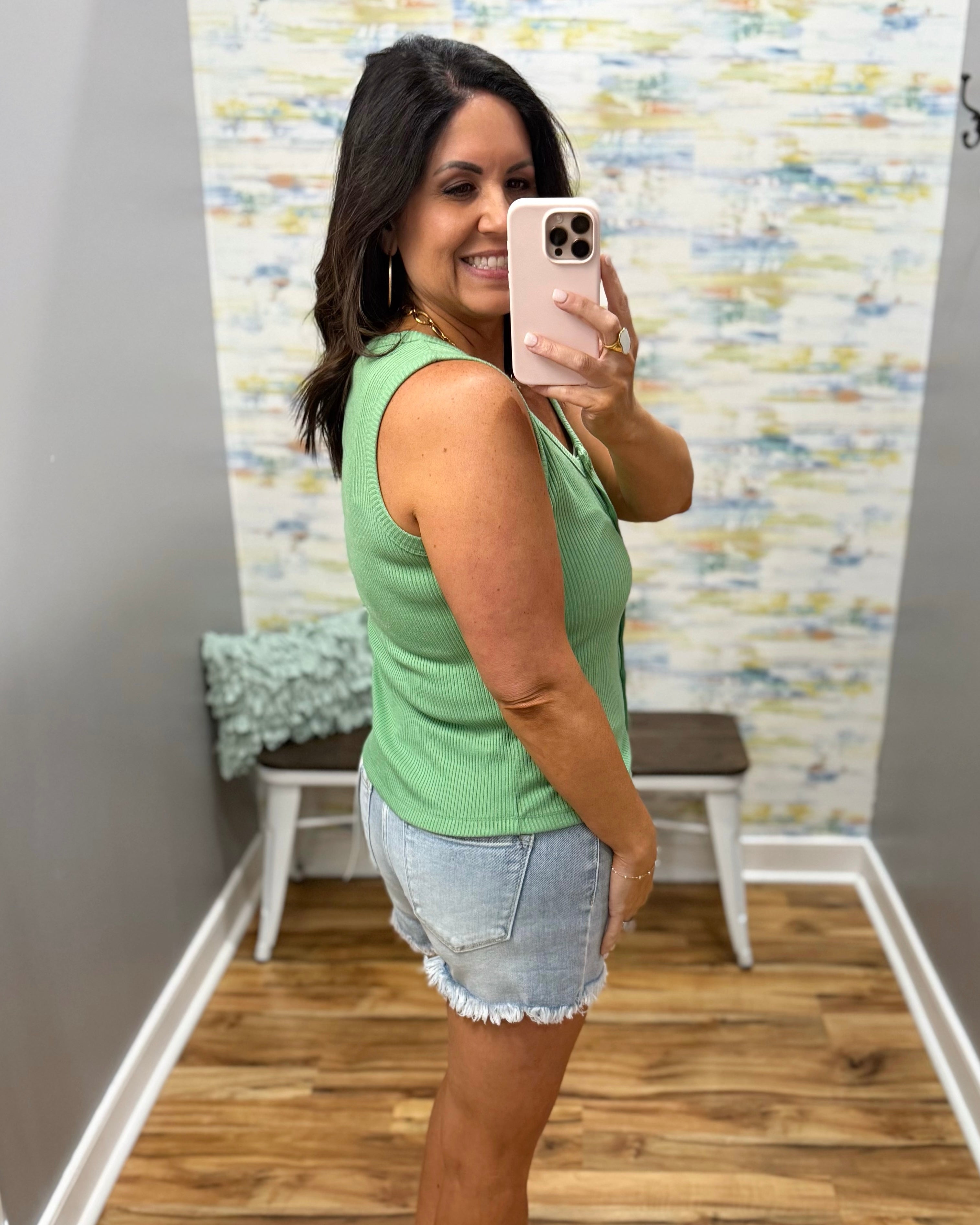 Woman taking a mirror selfie wearing a green sleeveless top and denim shorts in a room with a colorful wall.