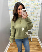 A woman takes a mirror selfie wearing a sage green knit sweater with white star patterns, paired with blue jeans. She is standing in a fitting room with a wood floor and a colorful abstract wall art piece in the background.