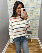 A woman takes a mirror selfie wearing a white knit sweater with horizontal stripes in rust red, olive green, and navy blue, paired with blue jeans.