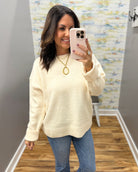 A woman takes a mirror selfie wearing a cream-colored knit sweater and blue jeans in a fitting room with a wood floor and abstract wall art.