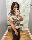 A woman takes a mirror selfie in a fitting room wearing a beige and black leopard print mock-neck top with ruffled sleeves and dark brown pants.