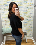 A woman takes a mirror selfie wearing a black short-sleeve knit top and blue jeans in front of a patterned wallpapered wall.