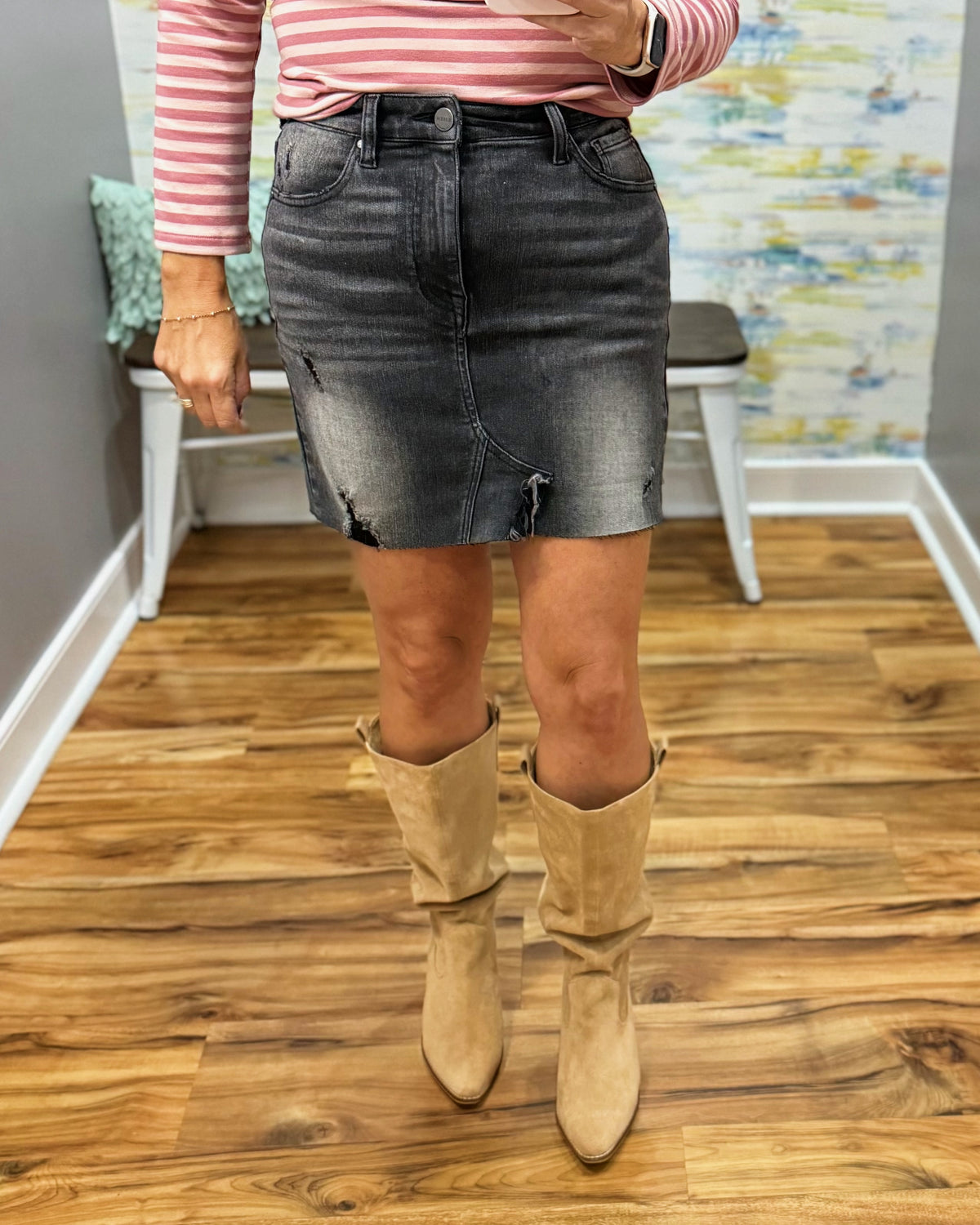 A person wearing a dark gray distressed denim mini skirt with ripped details, paired with a pink and white striped long-sleeve top and beige slouchy knee-high boots, standing on a wood floor.