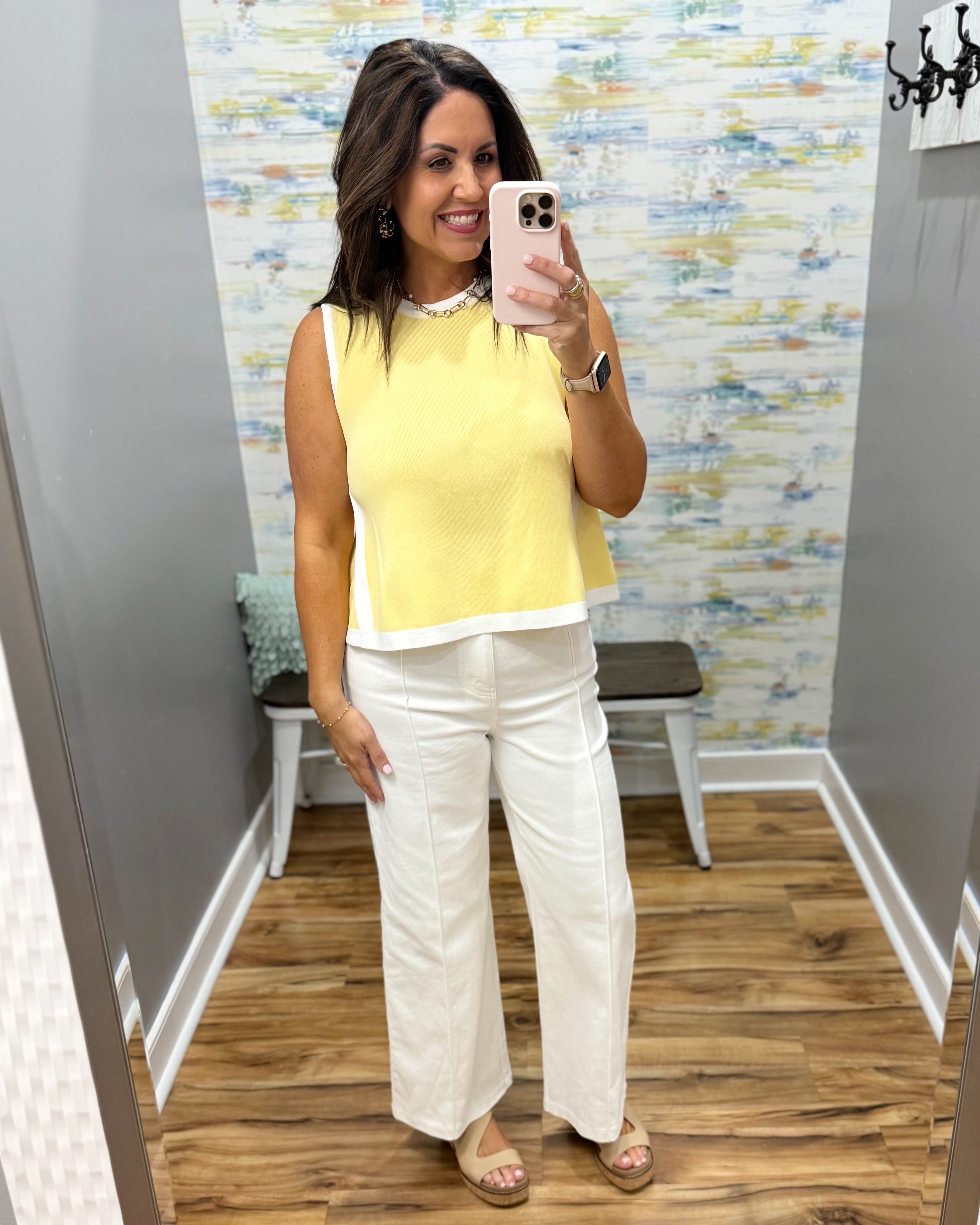 Woman taking a mirror selfie wearing a yellow top and white pants in a room with a patterned wall.