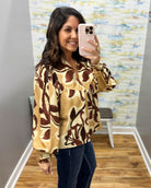 A woman takes a mirror selfie wearing a long-sleeve blouse with a brown and gold abstract botanical pattern, paired with dark blue jeans.