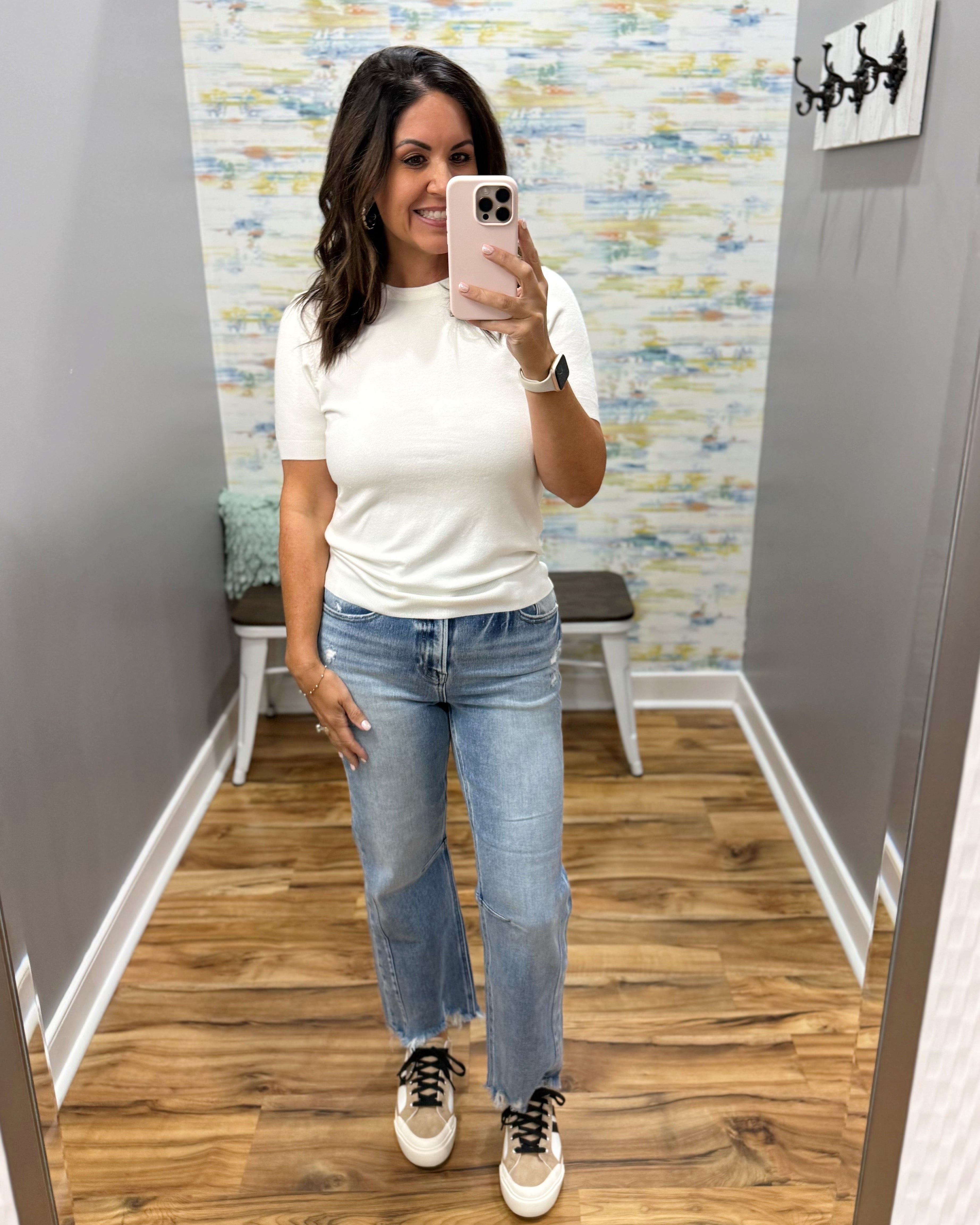 A woman takes a mirror selfie in a fitting room wearing a fitted white short-sleeve top, light-wash blue straight-leg jeans, and white sneakers with black laces.