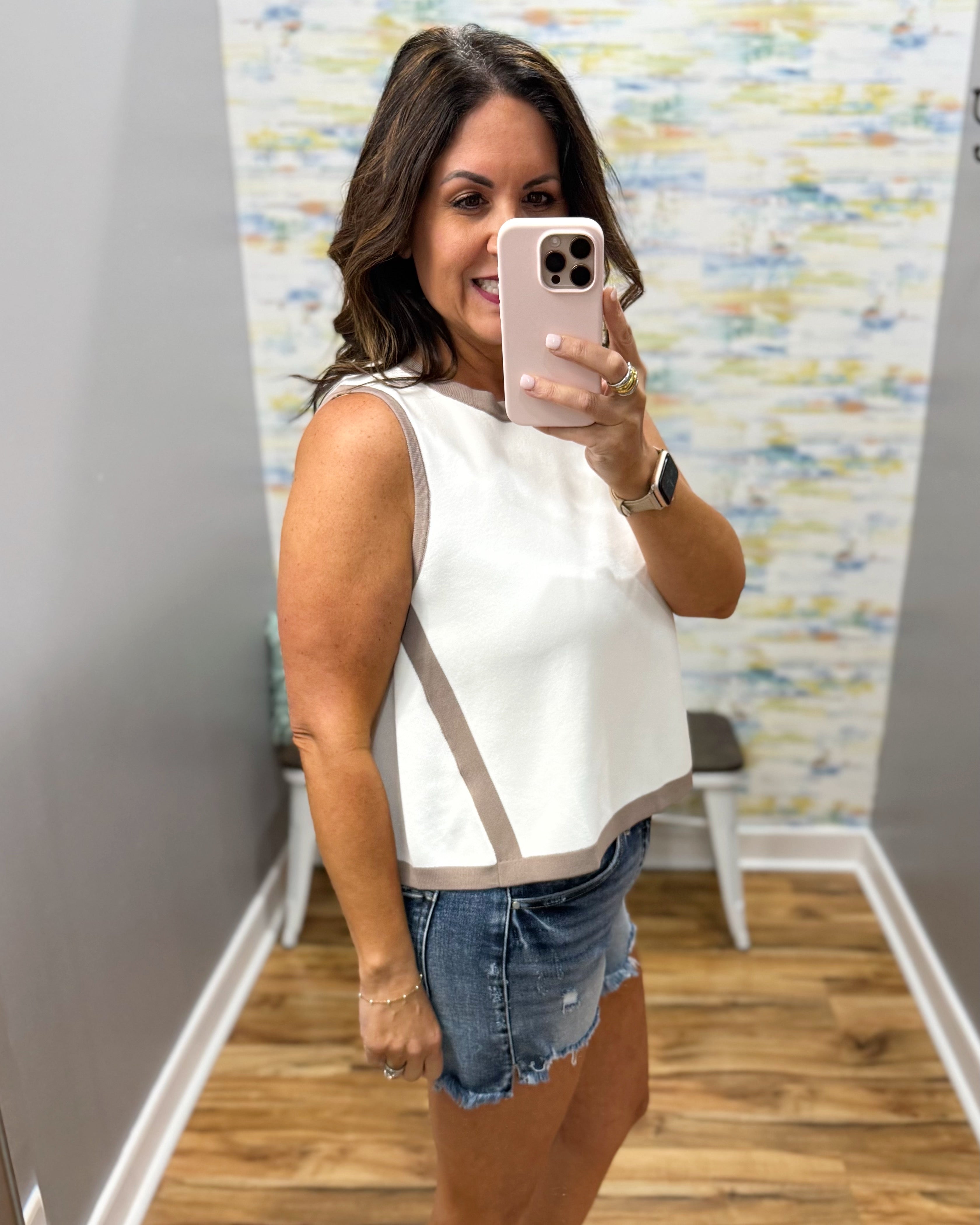 Woman taking a mirror selfie wearing a white sleeveless top and denim shorts in a room with a colorful wall.