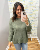 A woman takes a mirror selfie wearing a textured sage green long-sleeve knit top with a chest pocket, paired with light blue jeans.