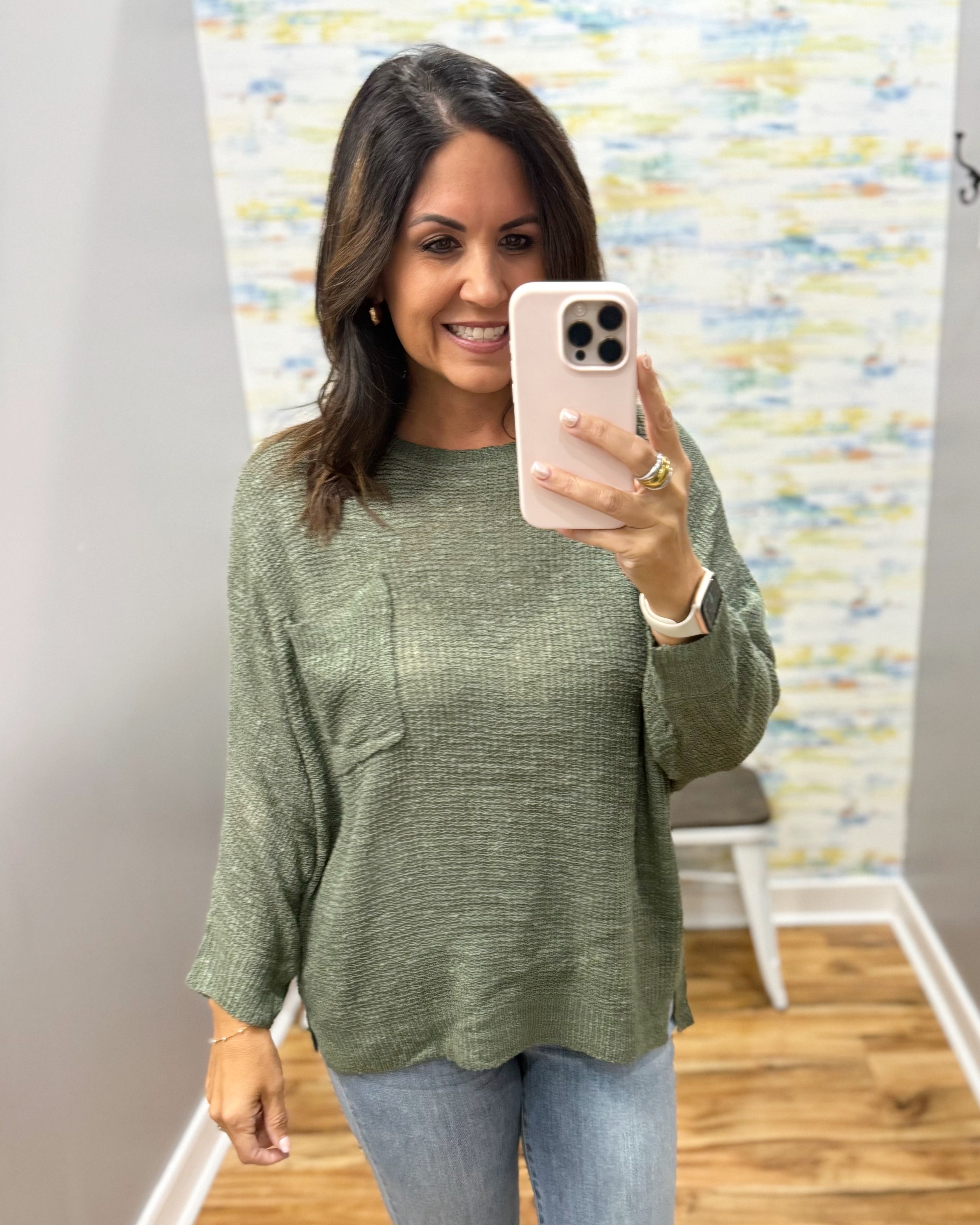 A woman takes a mirror selfie wearing a textured sage green long-sleeve knit top with a chest pocket, paired with light blue jeans.