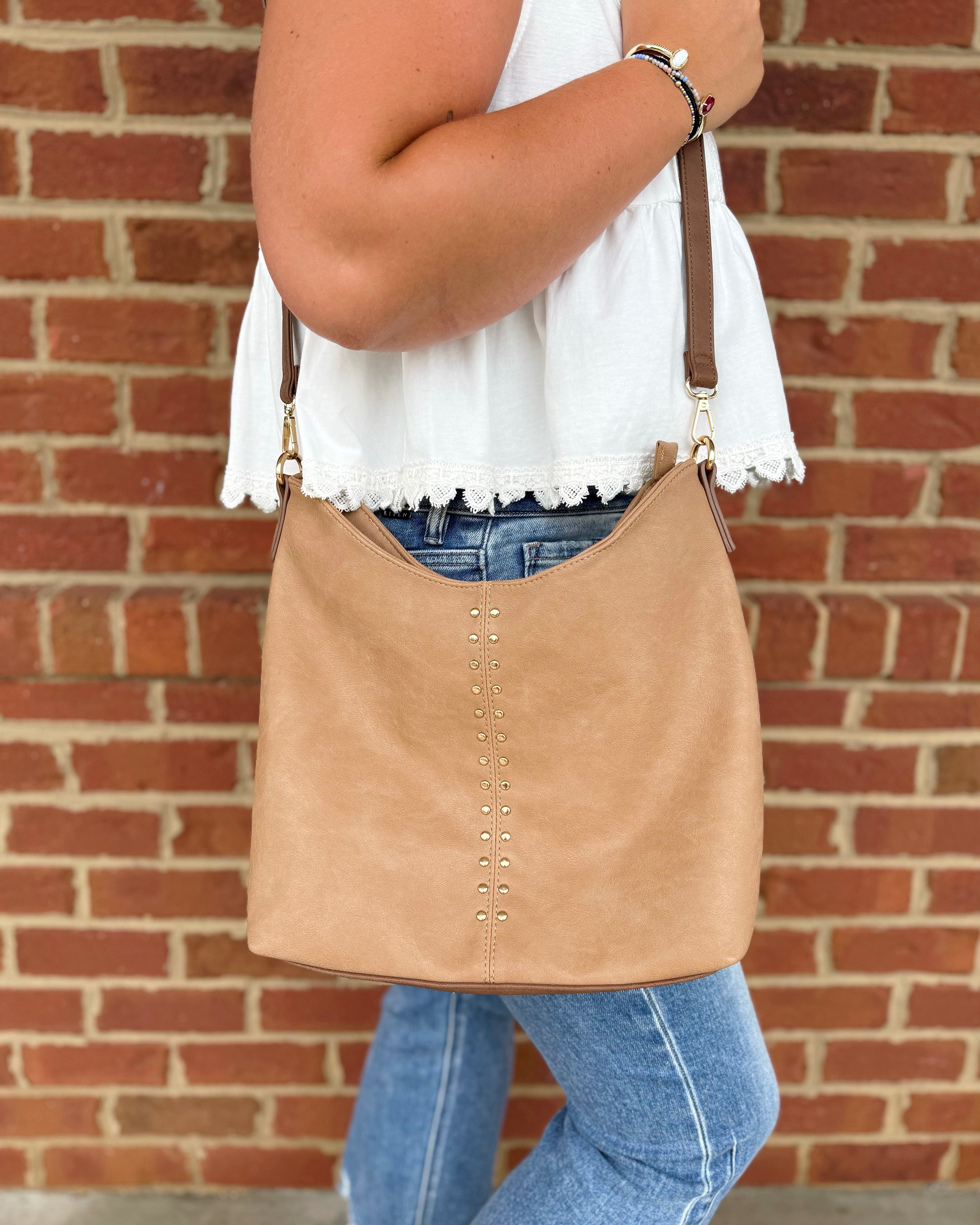 A tan suede crossbody bag with gold-tone hardware and a vertical row of silver studs, worn by a model in a white top and blue jeans standing against a red brick wall.