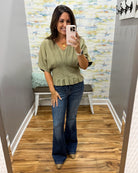 A woman takes a mirror selfie in a fitting room wearing an olive green short-sleeve blouse with a ruffled hem and dark blue flare jeans.