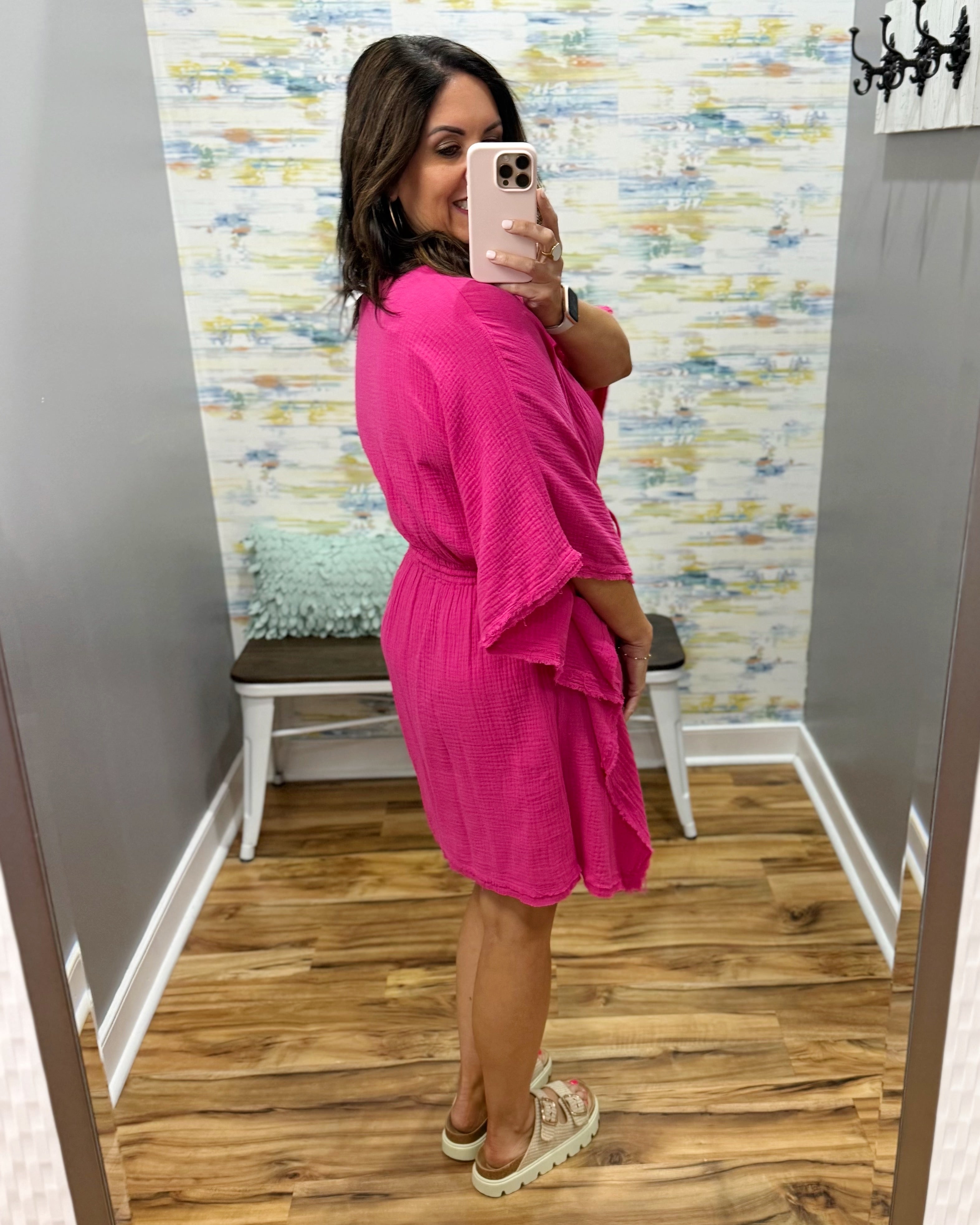 Woman wearing a pink dress in a fitting room with patterned wallpaper.