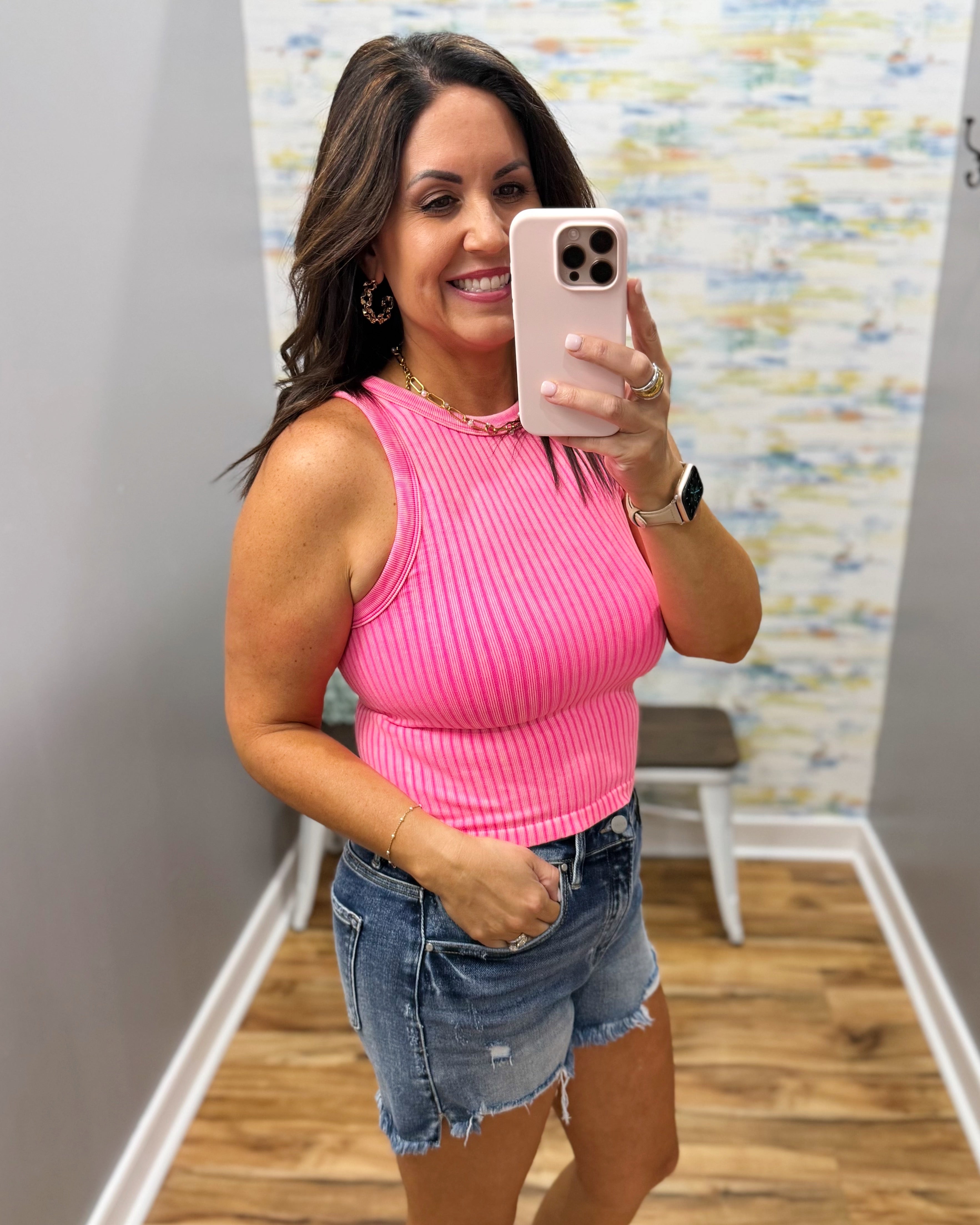 Woman taking a mirror selfie wearing a pink ribbed tank top and denim shorts.