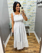 Woman wearing a white dress with a patterned wall and wooden floor in the background.