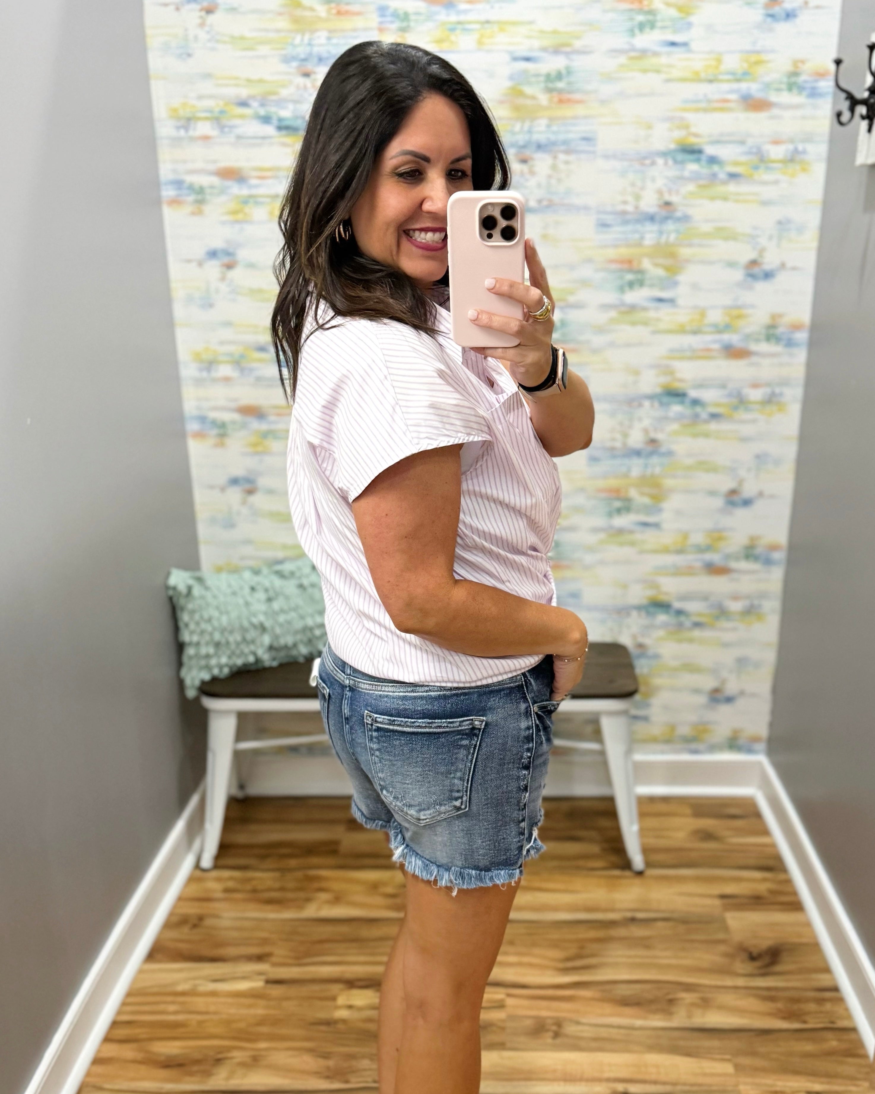 Woman taking a mirror selfie wearing a white shirt and denim shorts in a room with a textured wall.