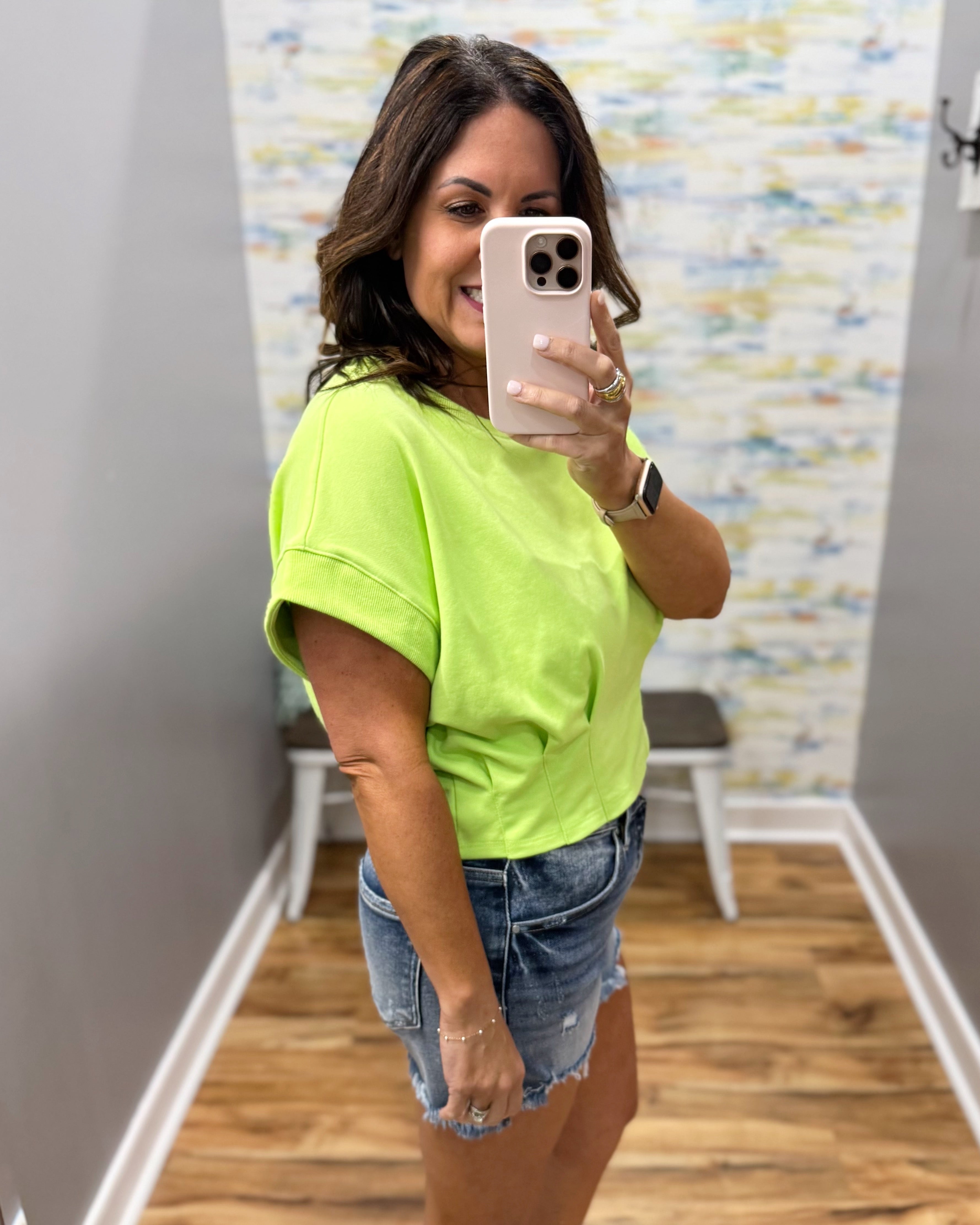Woman taking a mirror selfie wearing a bright green t-shirt and denim shorts in a room with colorful wall art.