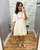 Woman taking a mirror selfie wearing a white dress with colorful embroidery in a room with a patterned wall.