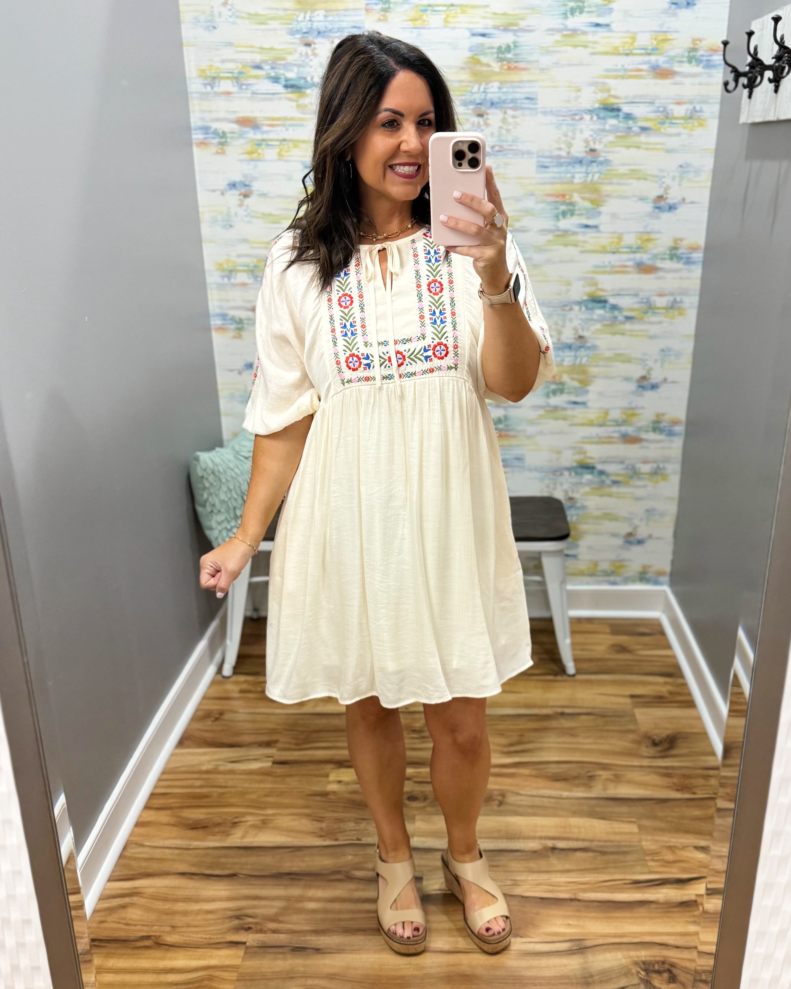 Woman taking a mirror selfie wearing a white dress with colorful embroidery in a room with a patterned wall.