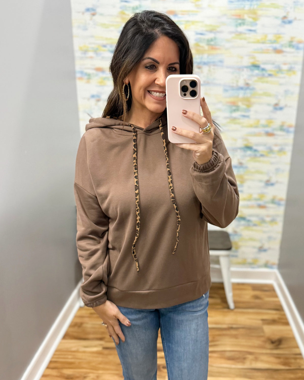 A woman takes a mirror selfie wearing a brown pullover hoodie with patterned drawstrings and blue jeans in an indoor room with a wood floor and abstract wall art.