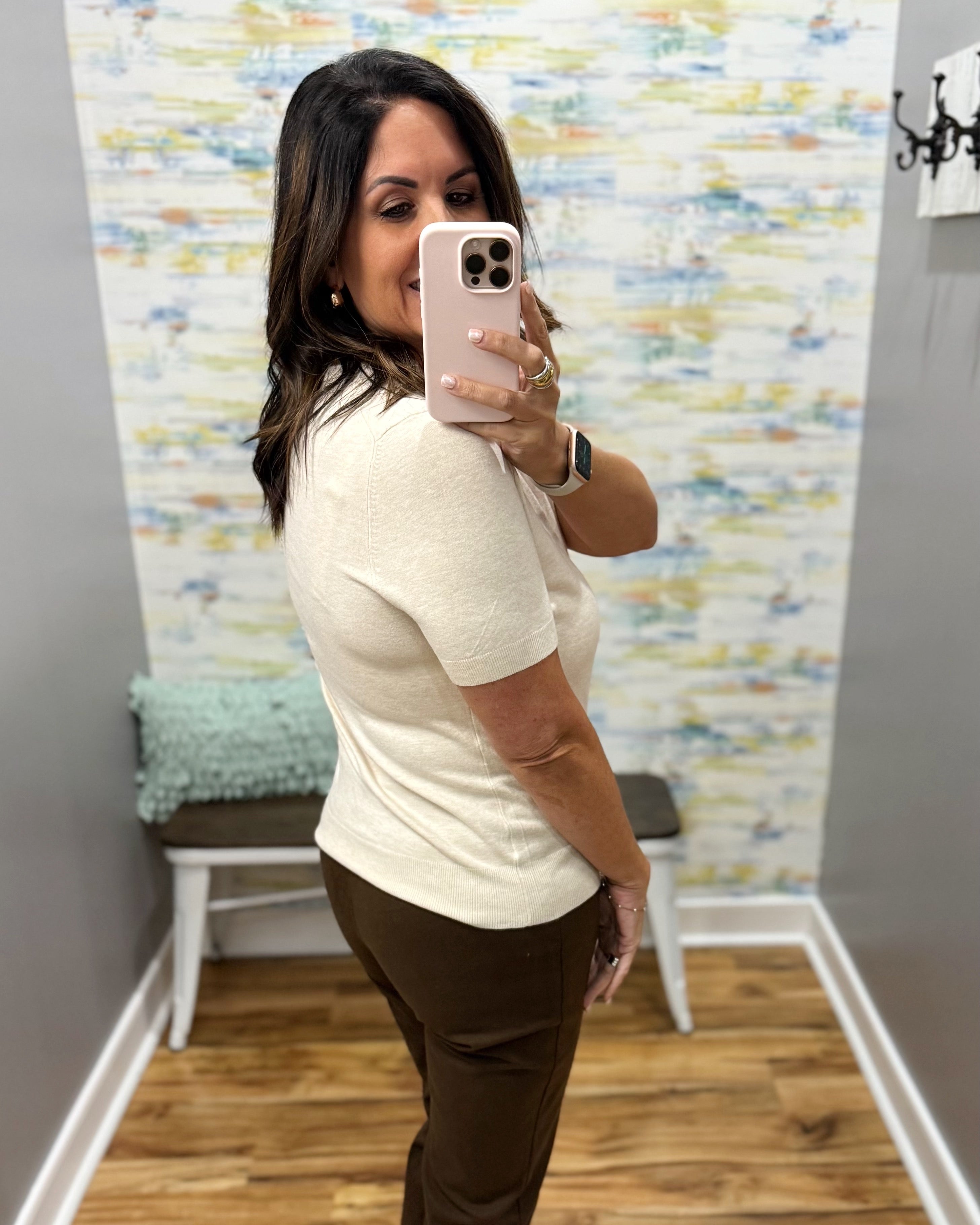 A woman takes a mirror selfie in a fitting room wearing a cream short-sleeve knit top and brown pants. The background features a colorful abstract patterned wallpaper, gray walls, and a wooden floor.